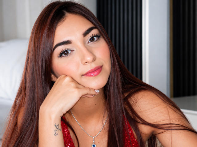 SalomeJoe with long brown hair, pink lips, and a tattoo on her hand, resting her chin on her hand, wearing a red sequined top, looking at the camera.