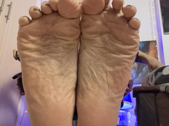 As solas dos pés de SarahRousen, enrugadas e levemente sujas, com unhas dos pés pintadas de verde, em uma foto tirada de baixo, mostrando seus pés em close. Seus dedos estão levemente curvados..