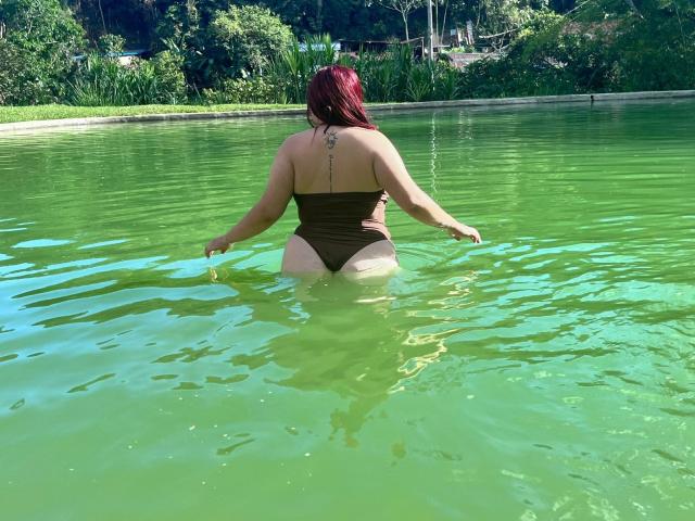 GeralGen con i capelli rossi, di spalle alla telecamera, in acqua verde, indossa un costume intero marrone, mostrando un tatuaggio sulla parte superiore della schiena, in piedi con le braccia aperte, con i glutei visibili..