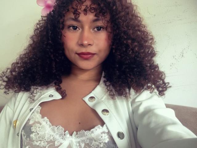 SofiaPassionne with curly brown hair, pink flower, and white open shirt, showing her lace-covered tits. She has medium brown skin, red lipstick, and a slight smile.