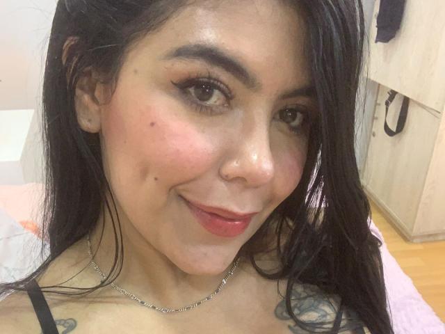 CatalinaReiyna with black hair, brown eyes, and red lips, smiling at the camera, showing a small nose boogey and several moles on her face, with a necklace.