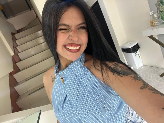 NyraVanel with long black hair, smiling with eyes squinted, wearing a blue striped halter top, showing tattoos on her right arm, standing in a white room.