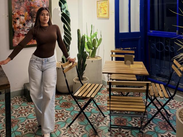 ThaliaAres with medium tits, brown long-sleeve top, light blue jeans, standing by wooden table, blue door, green plants, patterned floor, black chair, brown hair, white wall.