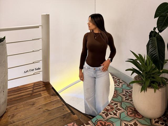 ThaliaAres, avec de longs cheveux bruns, un haut marron ajusté, un jean bleu taille haute, debout près d'un escalier en bois, une plante verte en pot, un mur blanc et un sol à motifs, regardant sur le côté..