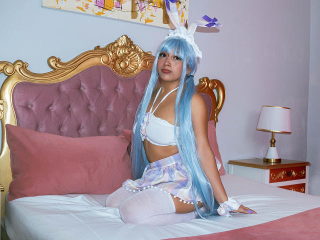 AilenRosse with blue hair, white lingerie, and pastel skirt sits on a pink bed, showing her tits and ass, wearing white thigh-highs and bunny ears.