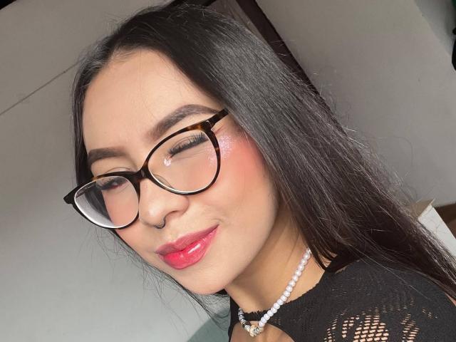 ChloeMoretti has shiny pink lips, black glasses, and a pearl necklace. She's smiling with her eyes closed, showing off her cute nose ring. Her ass is not visible.