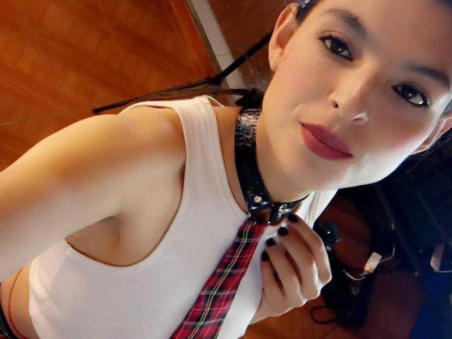 MerriethVelvet with light skin, dark hair in a ponytail, wearing a white tank top, black collar with a red plaid tie, holding the tie, black nails, smiling.
