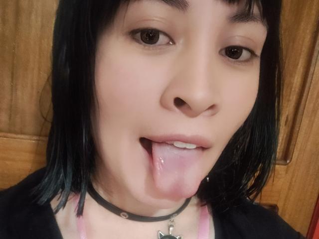El rostro de IssisDiosa, con cabello negro, ojos marrones y la lengua afuera, lleva un collar negro y un sujetador rosa, mostrando un pequeño piercing plateado en el labio..