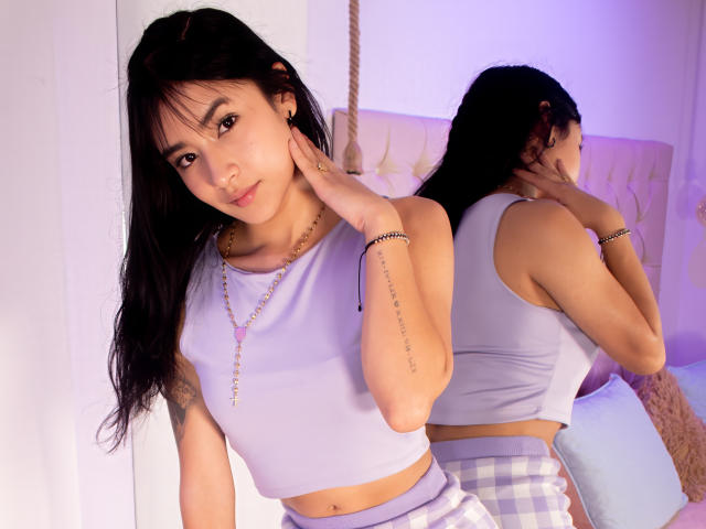 KatheLeclecr, com longos cabelos pretos, vestindo uma blusa cropped roxa clara e calças xadrez, toca em seu pescoço, refletindo em um espelho, mostrando seus pequenos seios e o braço tatuado..