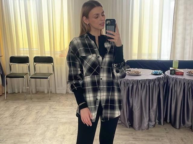Sanda Hayslett steht in einem Raum, trägt ein schwarz-weiß kariertes Hemd, schwarze Hosen und einen schwarzen Gürtel und macht ein Selfie mit ihrem Handy vor einem Spiegel..