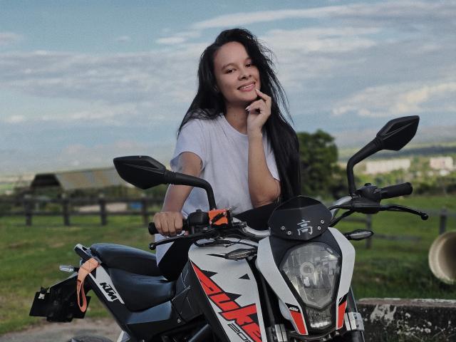 Yurimy with long black hair, white t-shirt, smiling, hand near mouth, sitting on a black and white motorcycle with red accents, outdoors, daytime, blue sky, and green grass.