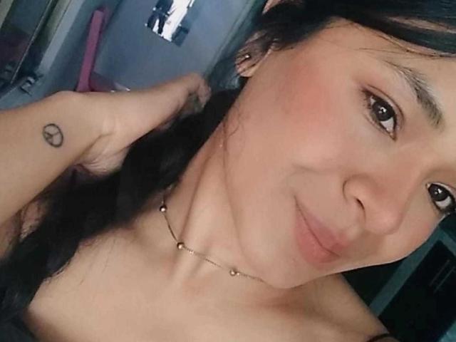 A selfie de AlissiaQueen mostrando sua pele bronzeada, olhos castanhos, cabelo preto em uma trança, um blush rosa suave, uma pequena tatuagem em formato de coração no pulso esquerdo e um colar delicado..