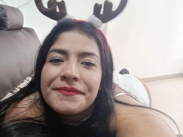 ArianithaXox with black hair, wearing reindeer antlers, smiling, red lipstick, lying on a brown couch, looking at the camera, showing her bare shoulders and upper chest.