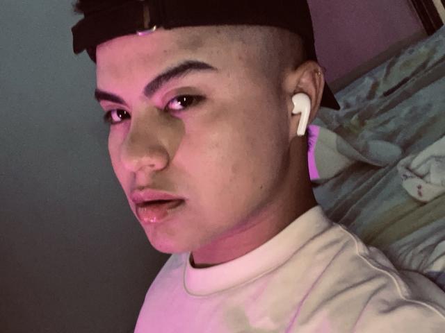 Thiago Ariass com um lado da cabeça raspado, boné preto, fones de ouvido brancos, luz rosa-roxo, camisa branca, lábios ligeiramente entreabertos e uma expressão séria, mostrando um piercing no nariz..