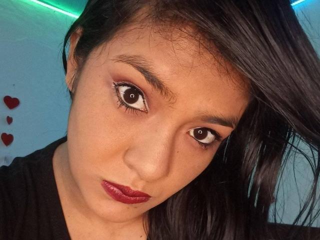 Il viso di ArianithaXox, occhi marroni, rossetto rosso, capelli neri e lisci, camicia nera, guarda la fotocamera, leggero inclinazione del capo, carina, sexy, incarnato medio, sopracciglia delicate, naso piccolo, espressione dolce..
