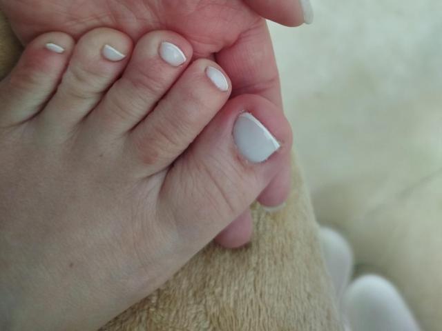 GladisBelle's hand with white-painted nails, holding her pinky to thumb, showing a slightly chipped nail on the pinky. Soft beige fabric underneath.