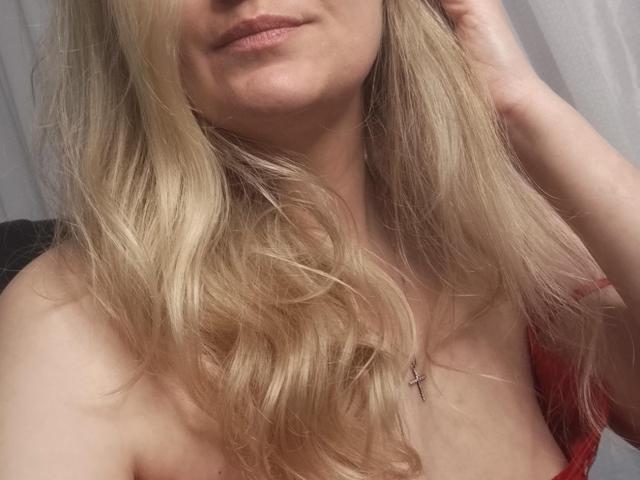 I capelli biondi e ondulati di GladisBelle, le labbra leggermente dischiuse, la pelle chiara e una croce al collo, con un abito rosso che lascia scoperte le spalle e il décolleté..