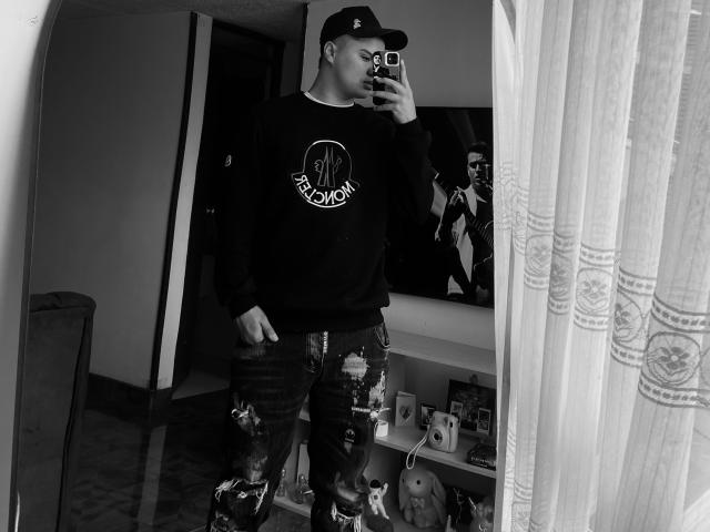 ThiagoAriass mit einer Baseballkappe, einem schwarzen Sweatshirt und zerrissenen Jeans, macht ein Selfie vor einem Spiegel in einem Raum. Er hält ein Handy mit einer gemusterten Hülle und zeigt seinen kahlrasierten Kopf..