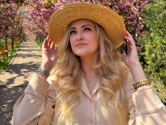 GladisBelle, avec ses cheveux blonds ondulés, portant un chapeau beige, une chemise beige et un bracelet doré, regarde vers le haut avec un léger sourire, les mains tenant le chapeau. Elle porte un collier avec une croix..