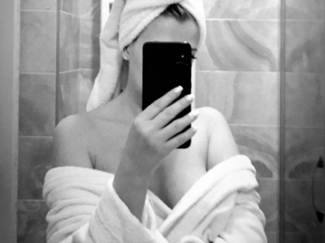 Black-and-white selfie of GladisBelle with a towel on her head, holding a phone over her face, wearing a robe that's off her shoulders, showing her small tits.