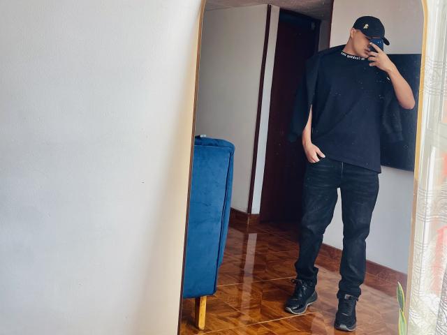ThiagoAriass, debout devant un miroir, portant une casquette noire, un t-shirt noir, un jean noir et des bottes noires, tenant un téléphone près de son visage, avec une chaise bleue..