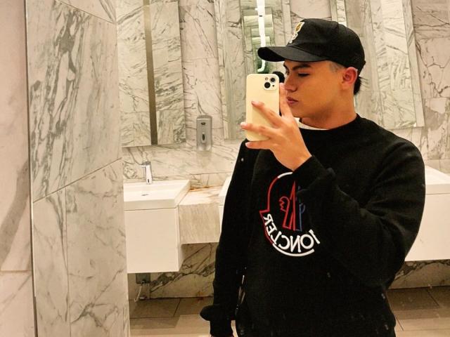ThiagoAriass, com um boné preto, moletom preto e um celular em um banheiro de mármore, tirando uma selfie no espelho, mostrando uma expressão de lábios emburrados. Ele tem um traseiro fofo e gordinho..