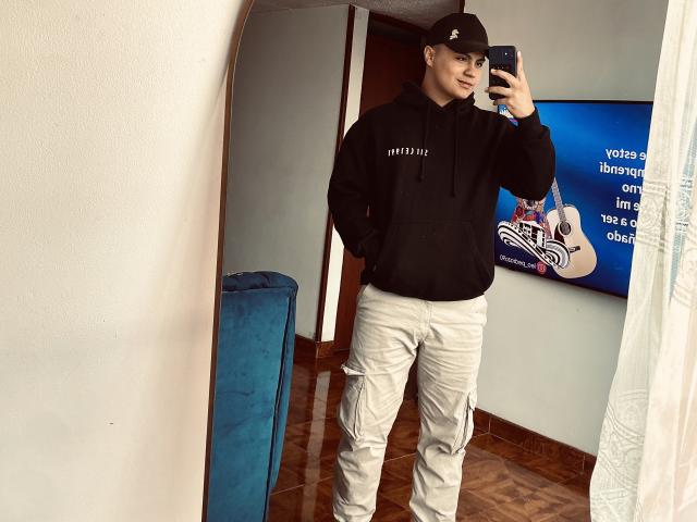 ThiagoAriass indossa una felpa nera e pantaloni cargo beige, porta un berretto nero ed è intento a scattare un selfie allo specchio con uno smartphone. Le sue mani sono nelle tasche..