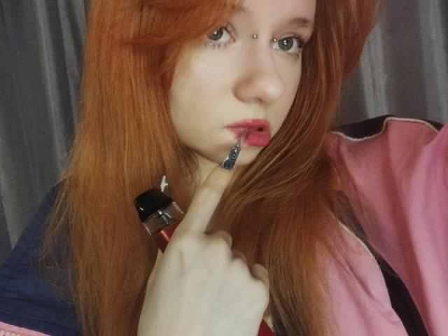 JamieLathrum with red hair, green eyes, and a nose piercing, biting a blue toothbrush, wearing a pink and blue jacket, touching her lips with her finger.