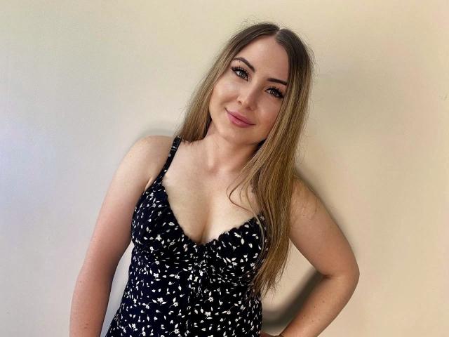 Une femme aux longs cheveux blonds, portant une robe noire avec des motifs blancs, a de petites seins, une peau claire et un léger sourire, et se tient devant un mur uni..