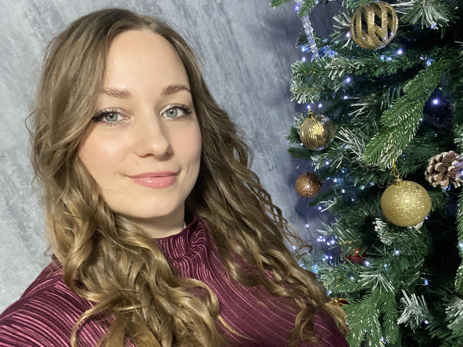 SueSuper with wavy brown hair, pink lips, and green eyes, wearing a red striped top, standing next to a decorated Christmas tree with gold ornaments and blue lights.