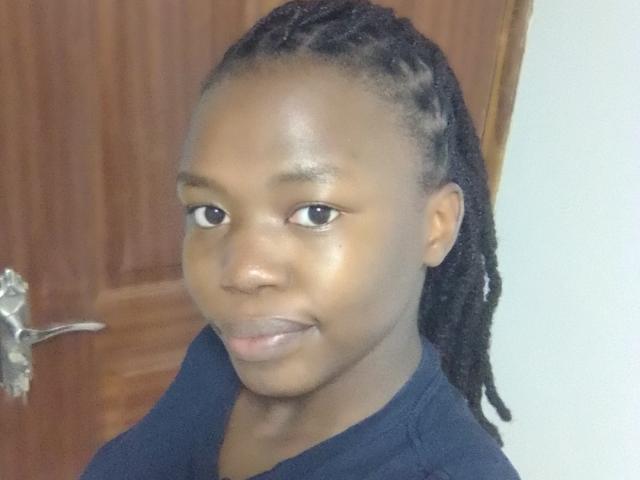 Yollah with dark skin, small nose, and long black dreadlocks, wearing a black shirt, smiling subtly, standing in front of a wooden door with a silver handle.