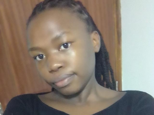 Yollah has dark skin, small lips, and braided hair. She's wearing a black shirt, looking slightly to the side with a subtle smile. Her ass is not visible.