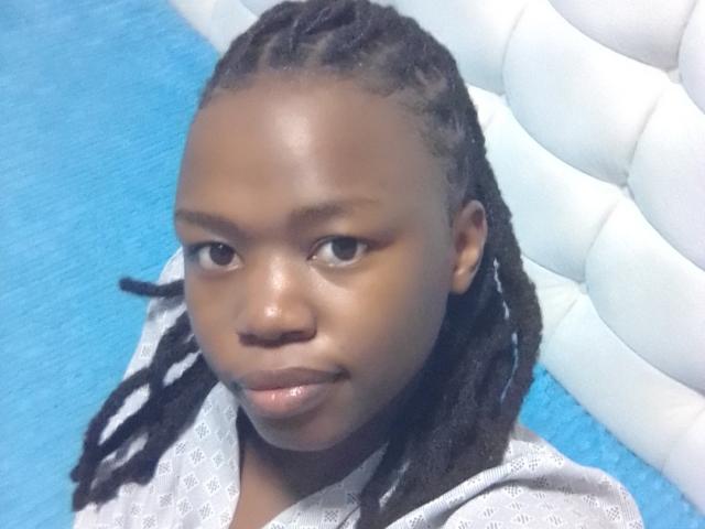 Yollah has dark brown skin, small shiny lips, and braided black hair. She's wearing a gray patterned shirt, looking straight at the camera with a slight smile.