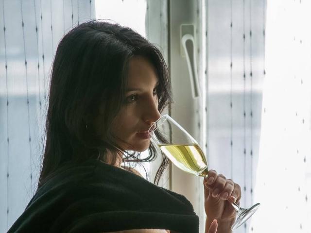 Sammy Hoffman, met donker haar, die een glas witte wijn vasthoudt, een zwarte sjaal draagt, naar de zijkant kijkt, zacht licht, serieuze uitdrukking, delicate vingers, zachte schaduw..
