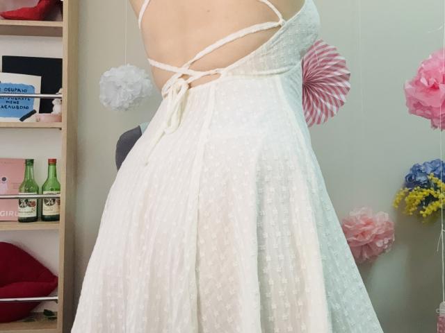 De witte, doorschijnende jurk van KarKutie met kleine stipjes, kruisende bandjes en een zwierige rok, die haar billen en rug laat zien, terwijl ze staat in een kamer met roze en witte papieren bloemen..