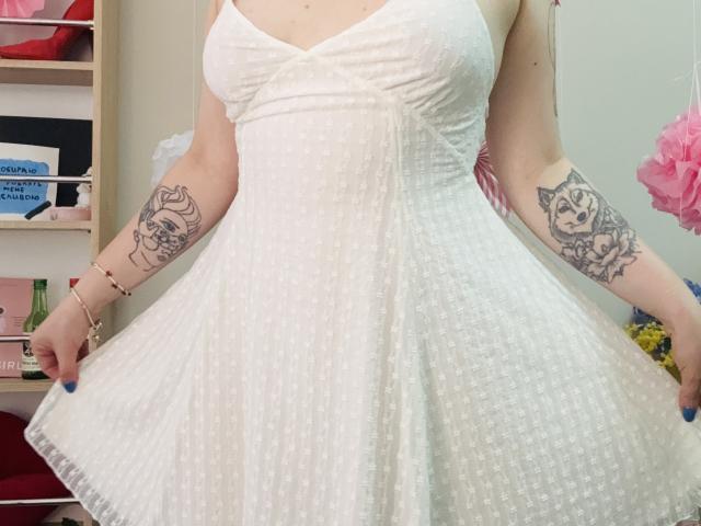 KarKutie draagt een witte, gestreepte, mouwloze jurk met zichtbare borsten, en houdt de rok uit. Ze heeft tatoeages op beide armen, blauwe nagels, en draagt een armband..
