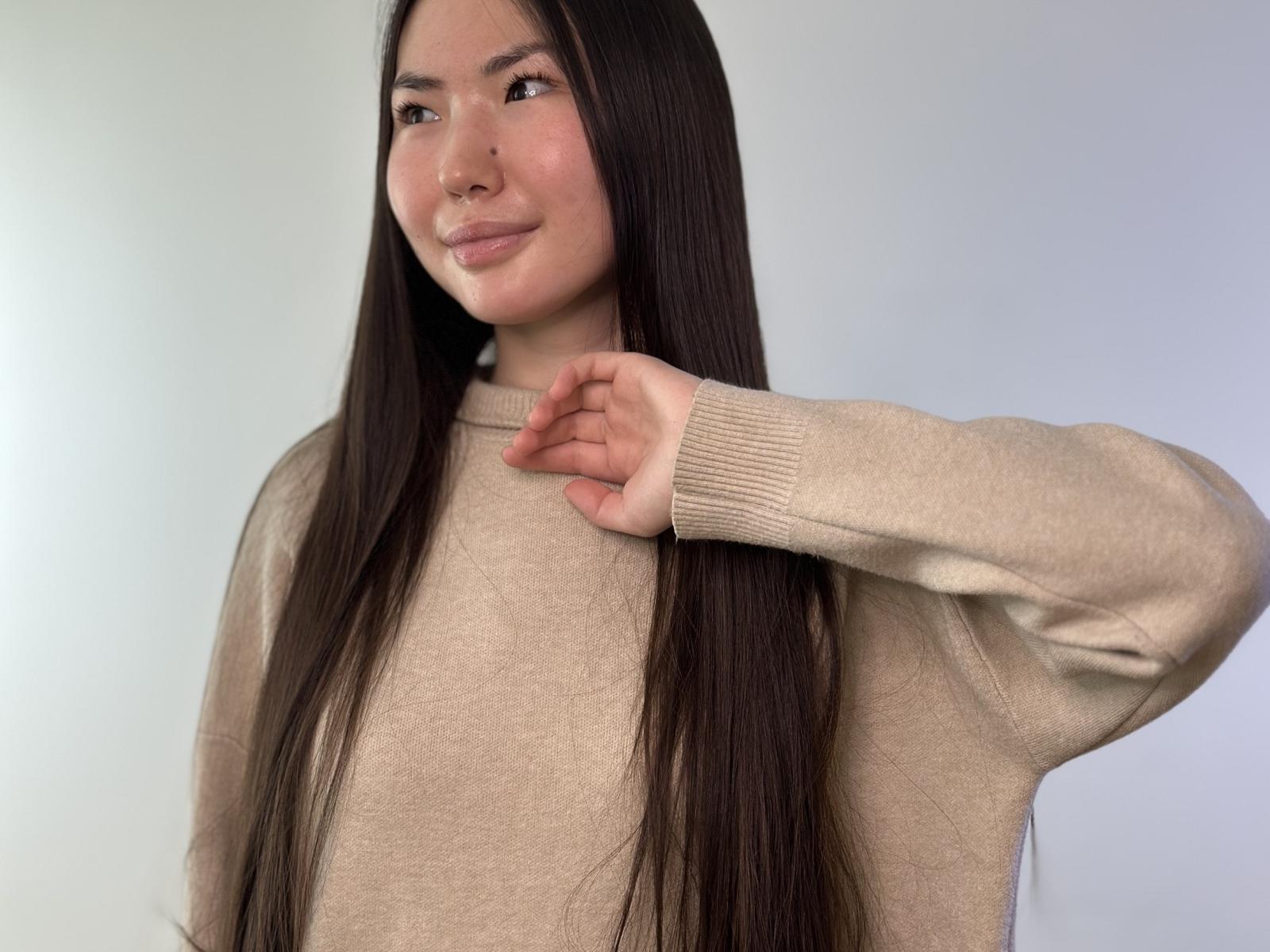 Adaline Russow, avec de longs cheveux foncés, un pull beige, un léger sourire, la main droite près du cou, une lumière douce, une peau claire, un maquillage discret, une expression douce, un style décontracté, un arrière-plan simple..