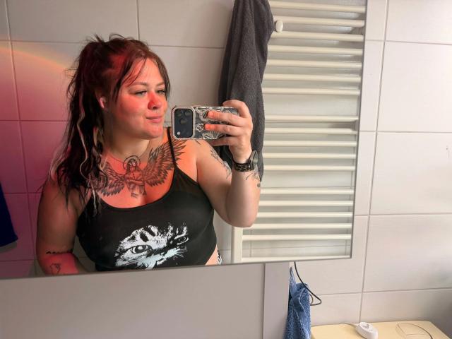 Prettyjosi, très sexy, porte un débardeur noir, laissant entrevoir ses seins, avec un tatouage sur la poitrine et des tatouages sur les bras, prenant un selfie devant un miroir dans une salle de bain. Une lumière rouge illumine sa joue..