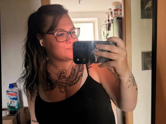 Prettyjosi-belle, avec des lunettes, un débardeur noir, un tatouage de hibou sur la poitrine, tenant un téléphone, selfie devant un miroir, cheveux bruns ondulés, tatouages sur le bras, une bouteille en arrière-plan, expression sérieuse, lumière douce..
