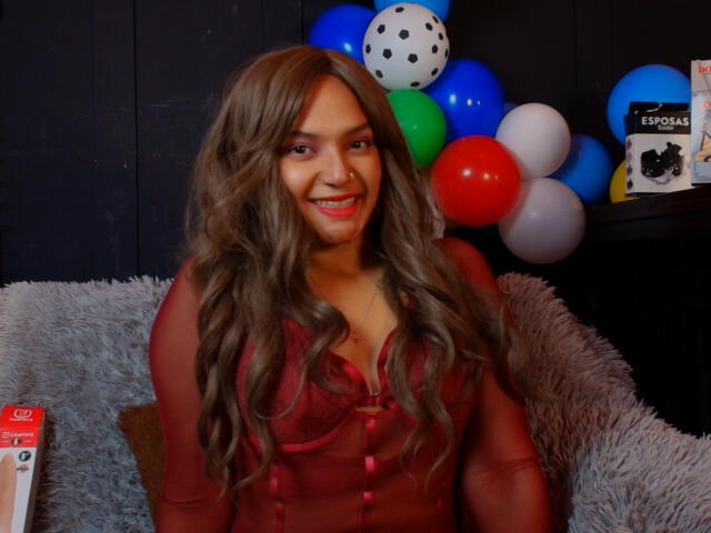 BrianaPratt with long brown wavy hair, red sheer top, showing her tits, smiling at camera, sitting on fluffy gray chair, colorful balloons and black bookshelf in background.