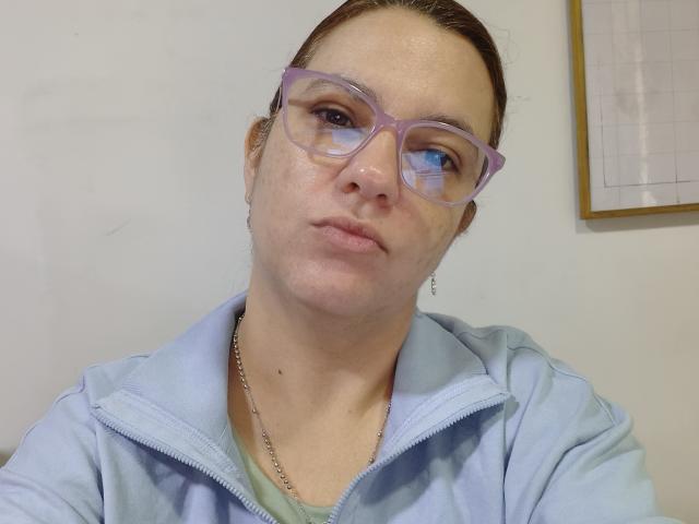 Amelia Wilson, com óculos roxos claros, lábios cheios e um leve bronzeado, vestindo uma jaqueta azul clara com zíper e uma corrente de prata, tem uma expressão neutra. Ela está olhando ligeiramente para a direita..