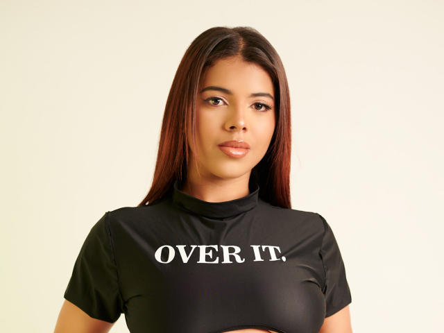 Una mujer de mediana edad con cabello castaño, piel media y labios brillantes, vestida con una camisa negra con la frase "OVER IT" en letras blancas, de pie frente a un fondo liso. Se pueden ver sus senos..
