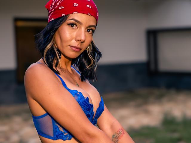 EvelinLuna met een blauwe kanten BH, bandana, golvend zwart haar, bruine huid, een kleine tatoeage op de linkerarm, subtiele make-up, staand, armen gekruist, kijkend naar de camera, buiten, avondlicht..