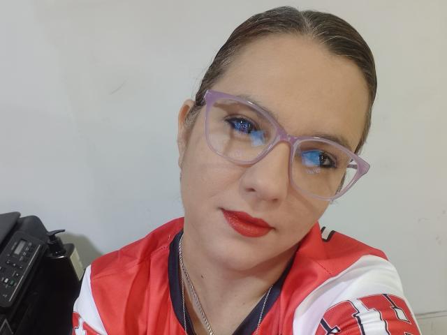 Amelia Wilson, com batom vermelho, óculos roxos e uma camisa esportiva vermelha e branca, exibindo suas nádegas com um colar de prata, cabelo castanho preso para trás, e uma parede branca neutra..