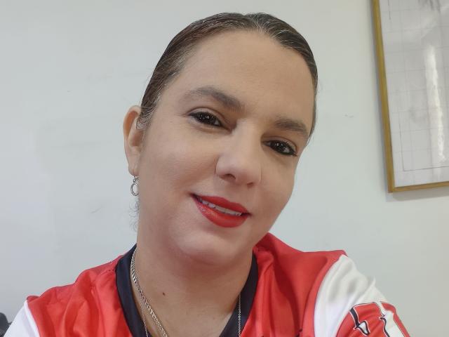 Amelia Wilson, con su cabello castaño peinado hacia atrás, lleva una camiseta deportiva roja y blanca, tiene lápiz labial rojo y delineador de ojos negro, y está sonriendo ligeramente..