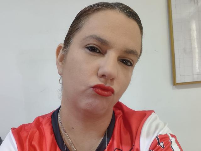 Amelia Wilson, con el cabello castaño peinado hacia atrás, lápiz labial rojo y una camiseta deportiva roja y blanca, frunciendo ligeramente los labios, llevando un collar de cadena y aros grandes..