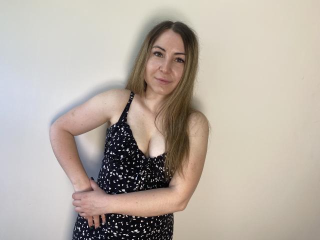 Aminas with long blonde hair, wearing a black, white-patterned dress, stands against a plain wall, her right hand on her hip, showing her small tits and a slight smile.