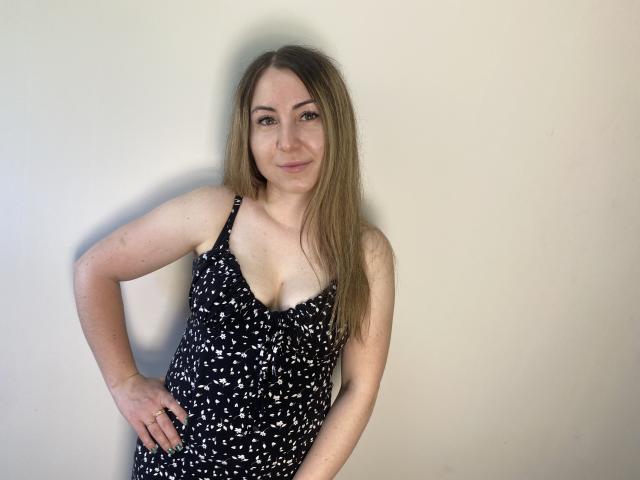 Aminas with medium tits, fair skin, and long brown hair, wearing a black and white patterned dress, standing against a plain wall, smiling slightly, hand on hip.