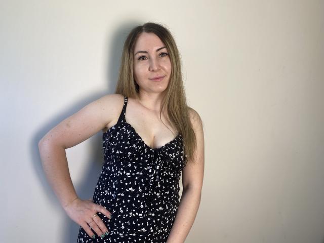 Une femme nommée Amina, avec de longs cheveux bruns, portant une robe noire et blanche à motifs, se tient contre un mur, une main sur la hanche, souriante, montrant sa petite poitrine et un joli visage..