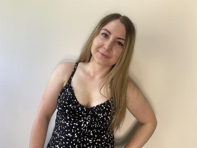 Aminas with long blonde hair, wearing a black and white patterned dress, showing small tits, leaning against a plain wall, smiling subtly.